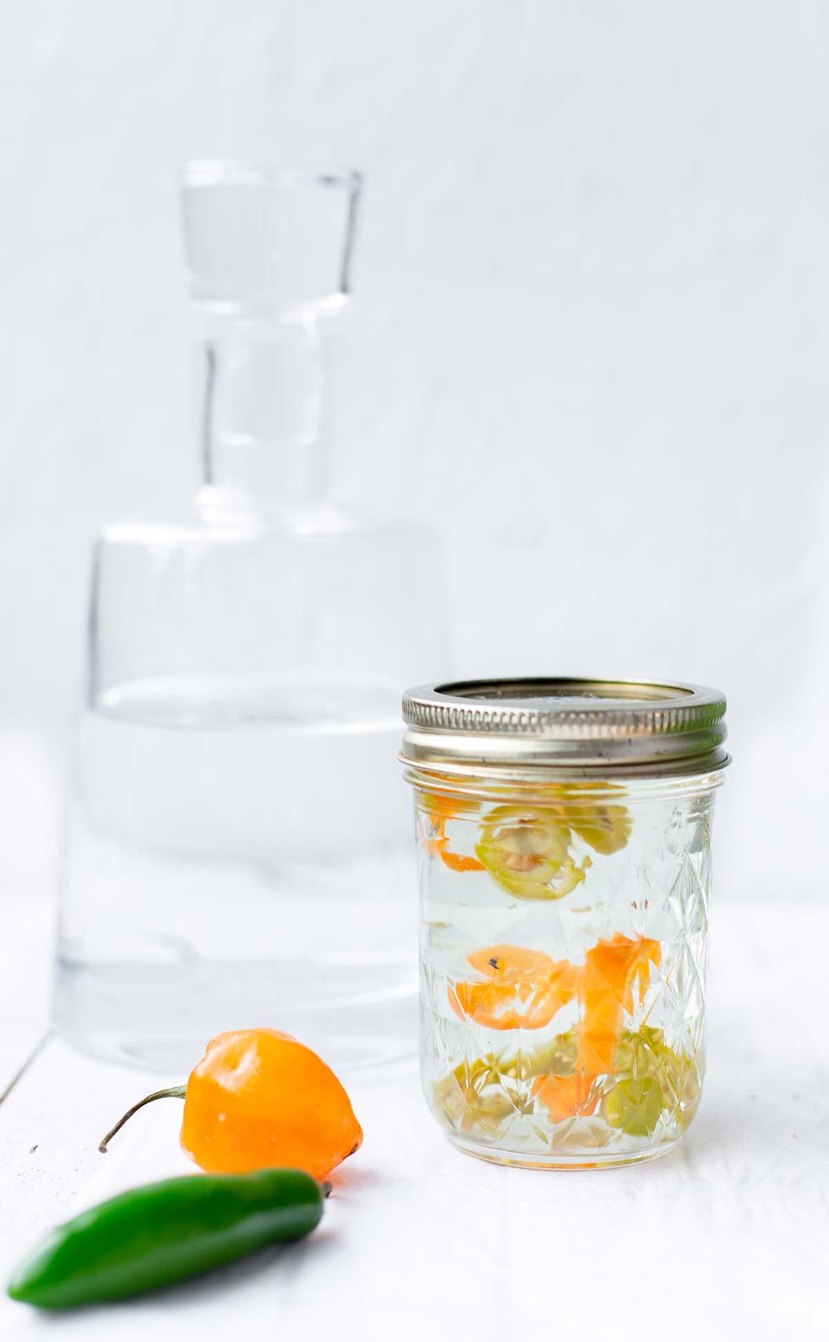 Large decanter of tequila with a smaller jar of habanero infused tequila in front of it and fresh chilies in the forefront.