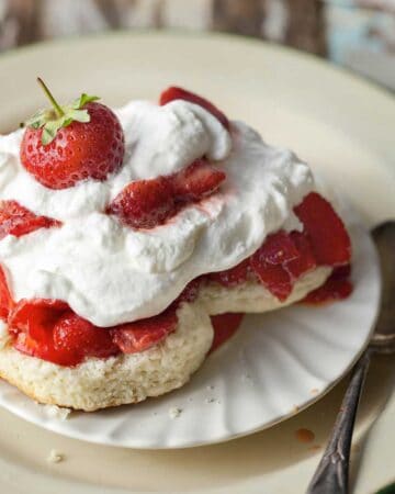 Open strawberry shortcake biscuit topped with juicy strawberries, freshly whipped cream and a few whole strawberries.