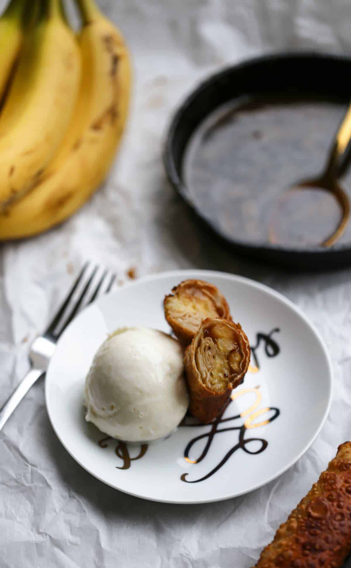 Banana spring rolls on a plate with ice cream and a skillet of warm caramel sauce in the background
