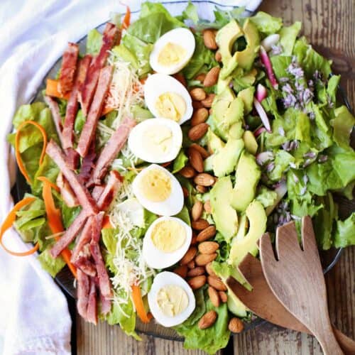A large round platter filled with greens, bacon strips, hard boiled egg halves, curly carrots, avocado slices and small purple chive blossom with wood salad servers on the side of the entree salad recipe.