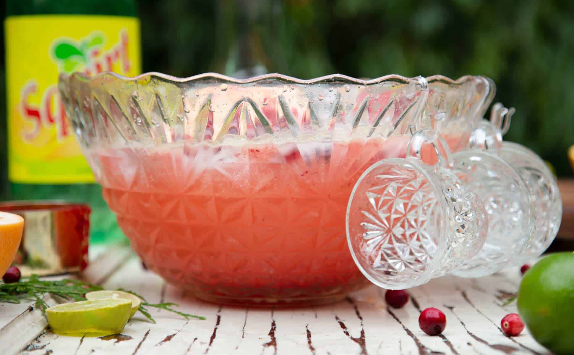 Side view of a vintage punch bowl filled with cranberry paloma punch with a bottle of grapefruit  soda in the background.