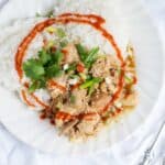 Bright white plate with shredded chicken teriyaki on a bed of rice with fresh herbs and hot sauce.