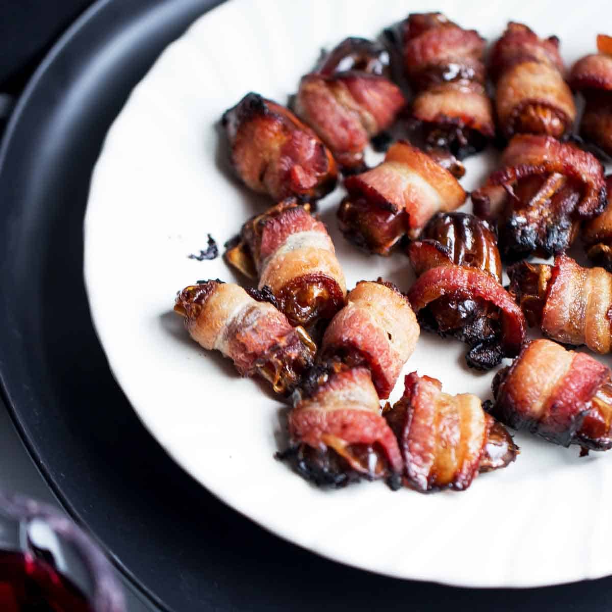 Crispy chewy browned bacon wrapped dates on a white plate with red wine.