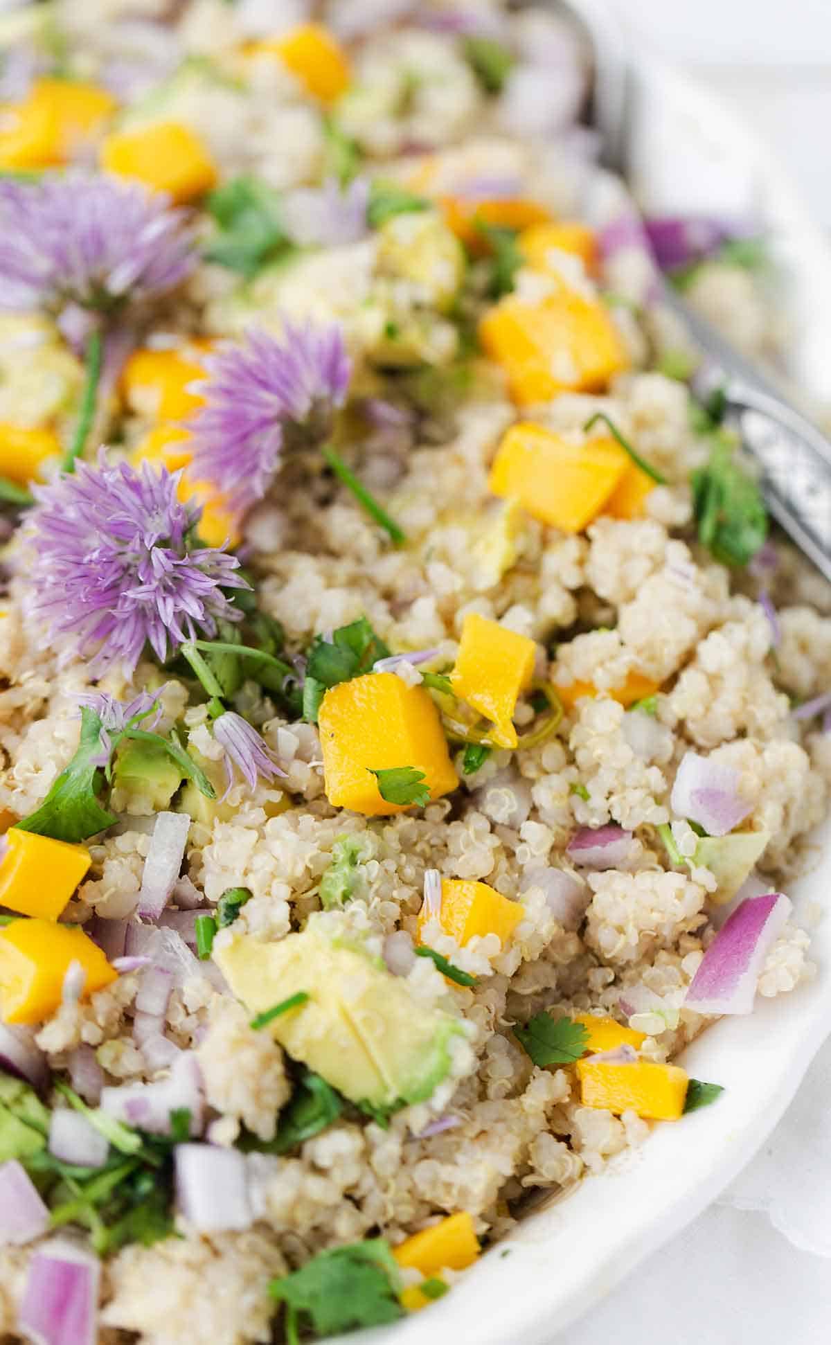 A platter of quinoa salad with mango pieces and purple chive blossoms on a white background.