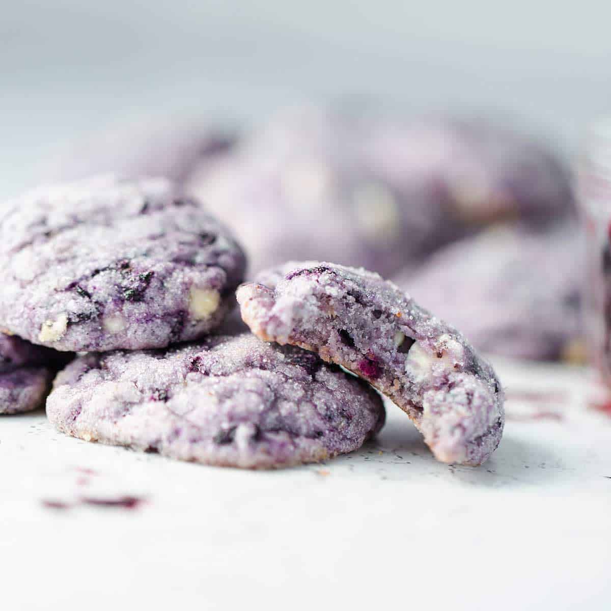 Fresh bright purple cookies with white chocolate stacked on a tray with frozen blueberries scattered around in addition to the blueberry cookie tray.