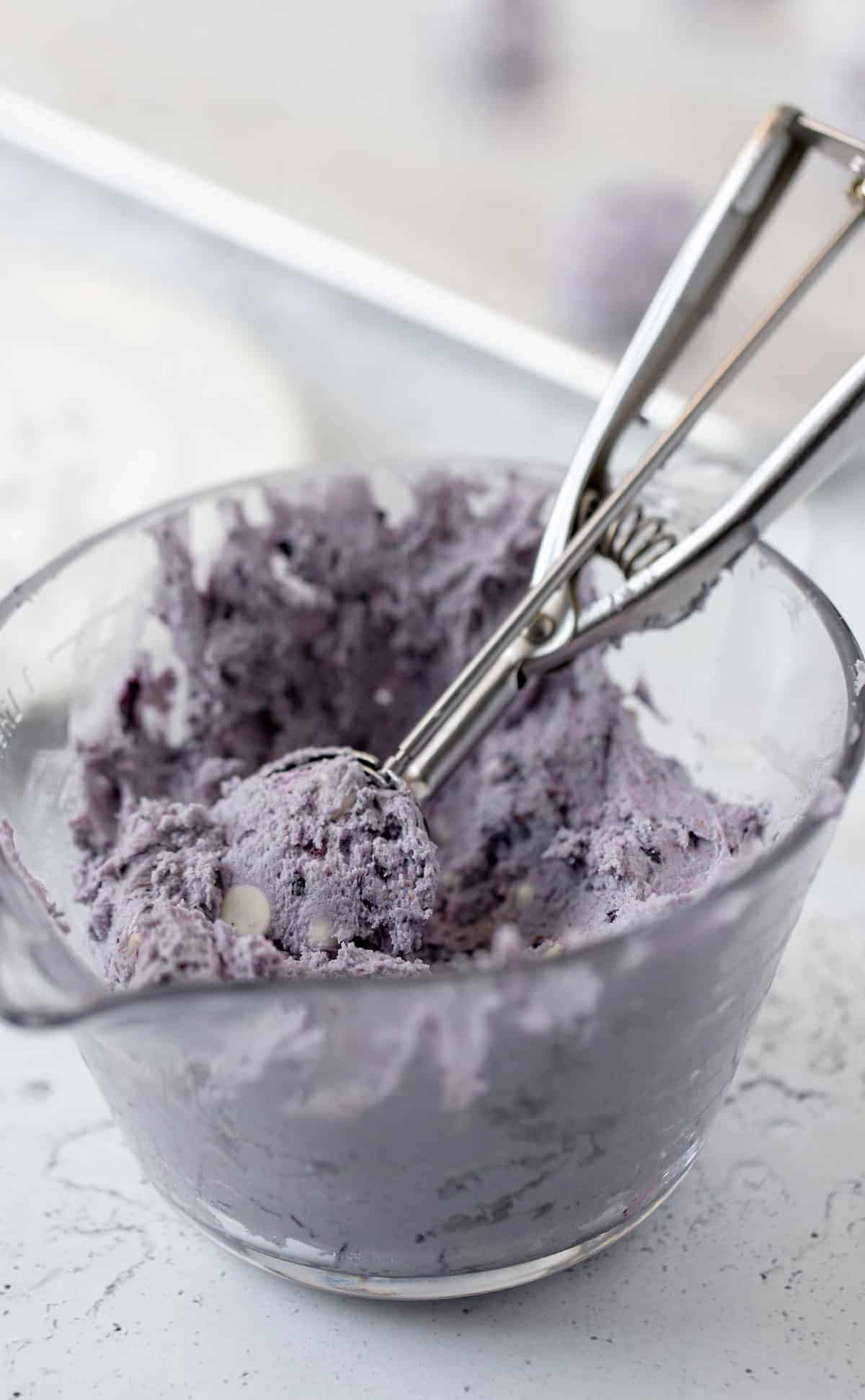 The soft blueberry cookie dough with white chocolate chips in a large glass measuring bowl with a cookie scoop sticking out