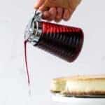 Blackberry sauce being poured from an old fashioned syrup dispenser over cheesecake.