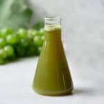 A beaker of naturally green grape juice with fresh grapes in the background.