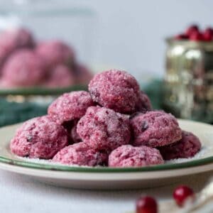 Cranberry cookies that are bright maroon red and covered with coarse sanding sugar to look like sparkling sugar plums are stacked high on a plate with fresh cranberries scattered around the foreground.