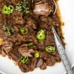 Tender shredded beef on a white platter garnished with sliced fresh shisto peppers and oregano blossoms.