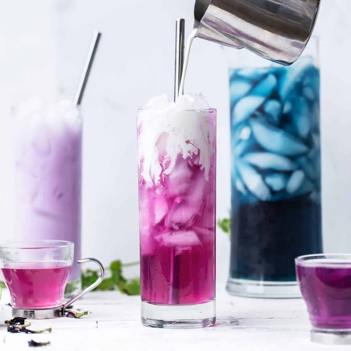 Color changing butterfly pea flower latte in shades of purple, pink and blue with cream drizzling down over the ice of the pink iced tea in front.
