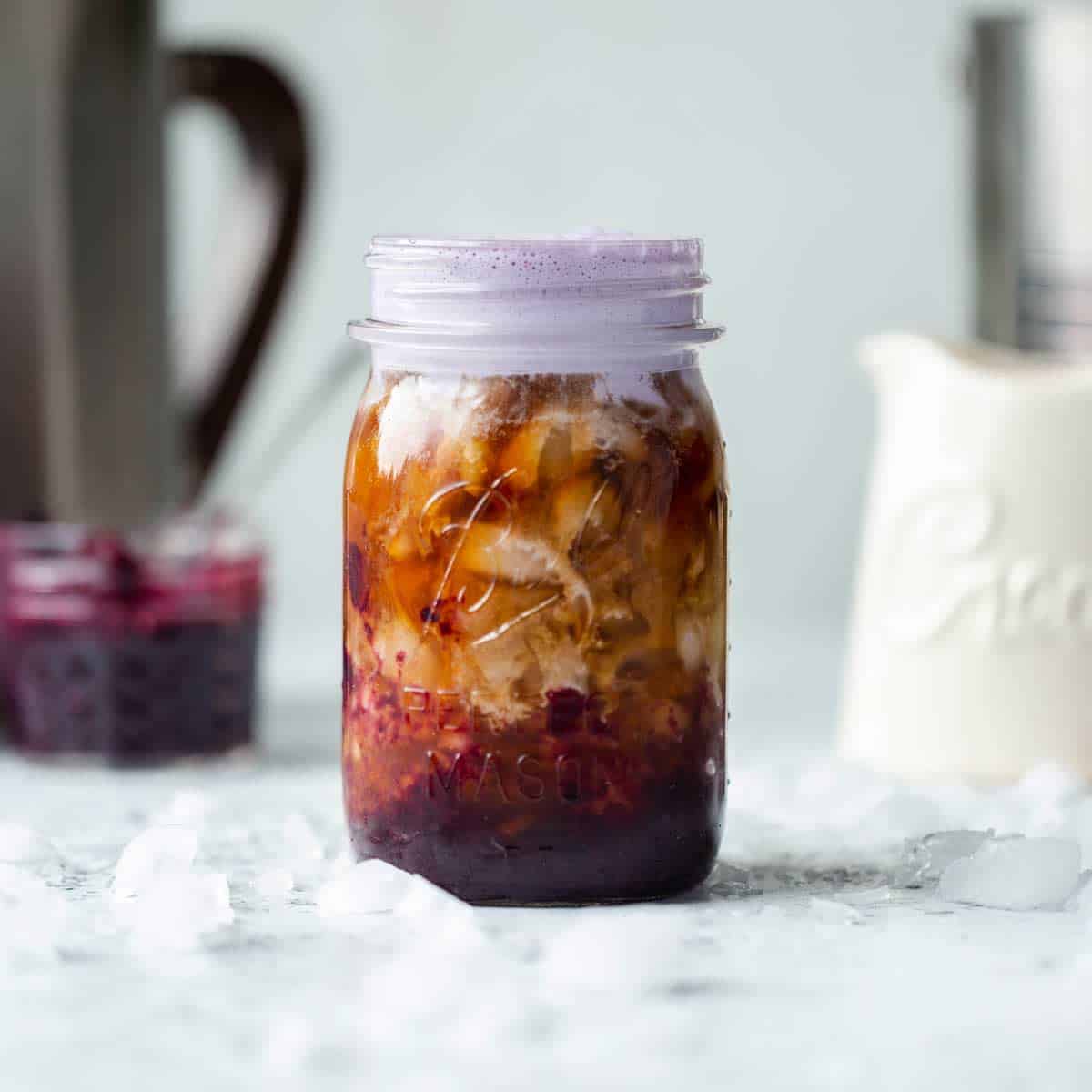 Blueberry coffee in a mason jar with purple blueberry cold foam on the top and cream, blueberry puree and coffee visible in the background.