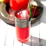 A pitcher and a glass of spicy pink strawberry lemonade in a glass with a chili pepper and strawberry garnish on rim.