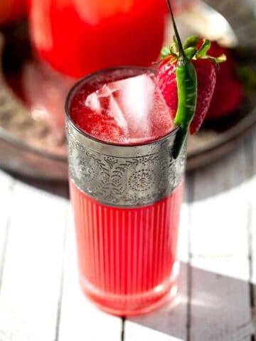 A pitcher and a glass of spicy pink strawberry lemonade in a glass with a chili pepper and strawberry garnish on rim.